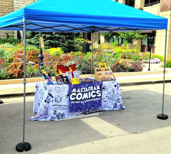 Photograph of Mastafran's table at Alley Art Fest 2026 in Aurora,IL. This picture showcases Mastafran's table and tent setup with his selection of merchandise including books, stickers, and tablemats.