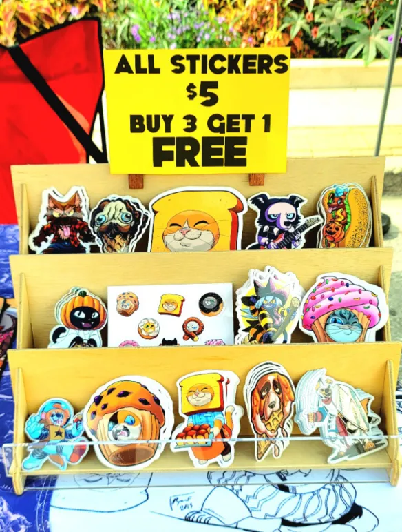 Photograph of Mastafran's table at Alley Art Fest 2026 in Aurora,IL. This picture showcases his selection of vinyl stickers for sale. All stickers are five dollars with a special to purchase three stickers and get an extra sticker for free.