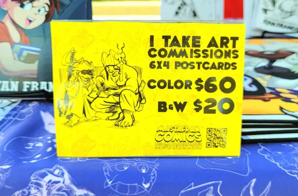Photograph of Mastafran's table at Alley Art Fest 2026 in Aurora,IL. Mastafran offers live art commissions on 6 inch by 4 inch post cards for sale. Color commissions with alcohol marker are $60, while black and white pen and ink commissions are $20.