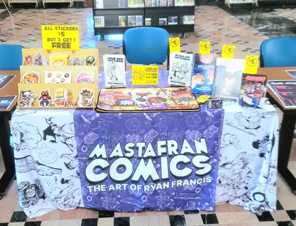 Photo of Mastafran's table at Fandom, Art, and Music Expo 2026, showing his selection of self-published comics, stickers, and tablecloths.